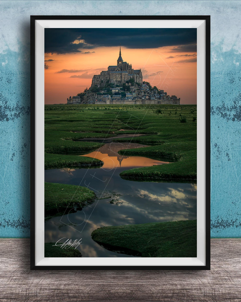 A framed photograph depicting the iconic Mont Saint-Michel, a medieval fortified island in Normandy, France, with its distinctive tower and spire against a vibrant sunset sky.