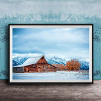 A large wooden barn with a sloping roof stands in a snowy field, with snow-capped mountains visible in the background.