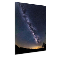 a stunning night sky with the Milky Way galaxy stretching across the frame, its bright white center contrasting against the dark blue backdrop. The silhouettes of trees can be seen in the distance, adding a sense of depth and scale to the scene.