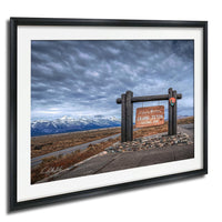 A framed photograph of a scenic landscape with a wooden sign reading "Grand Teton National Park" in the foreground, set against a backdrop of snow-capped mountains and a cloudy sky.