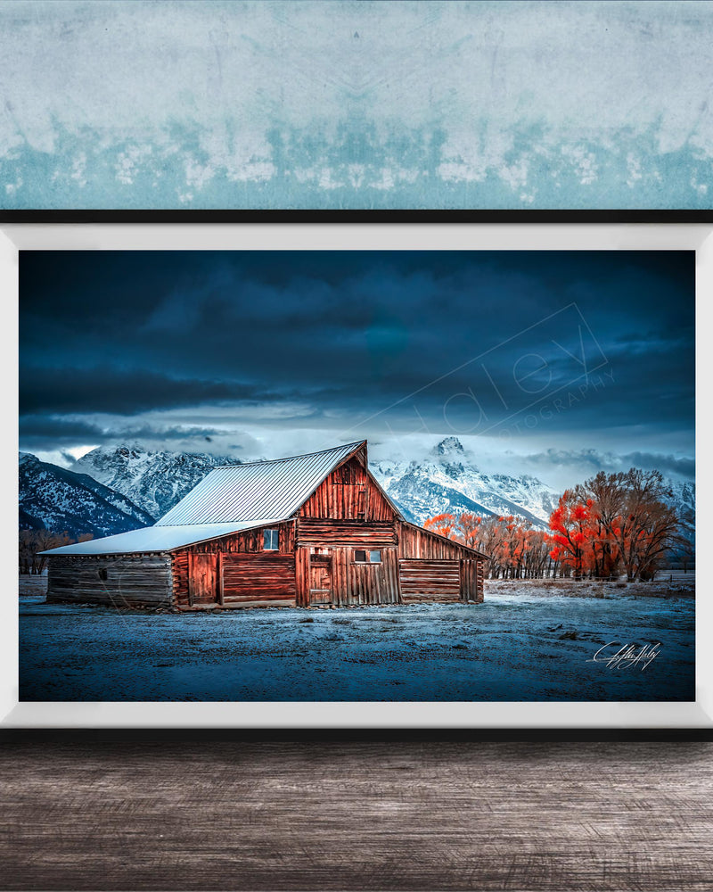 A rustic wooden barn stands in a snowy landscape, with a mountain range in the background under a dark, cloudy sky.