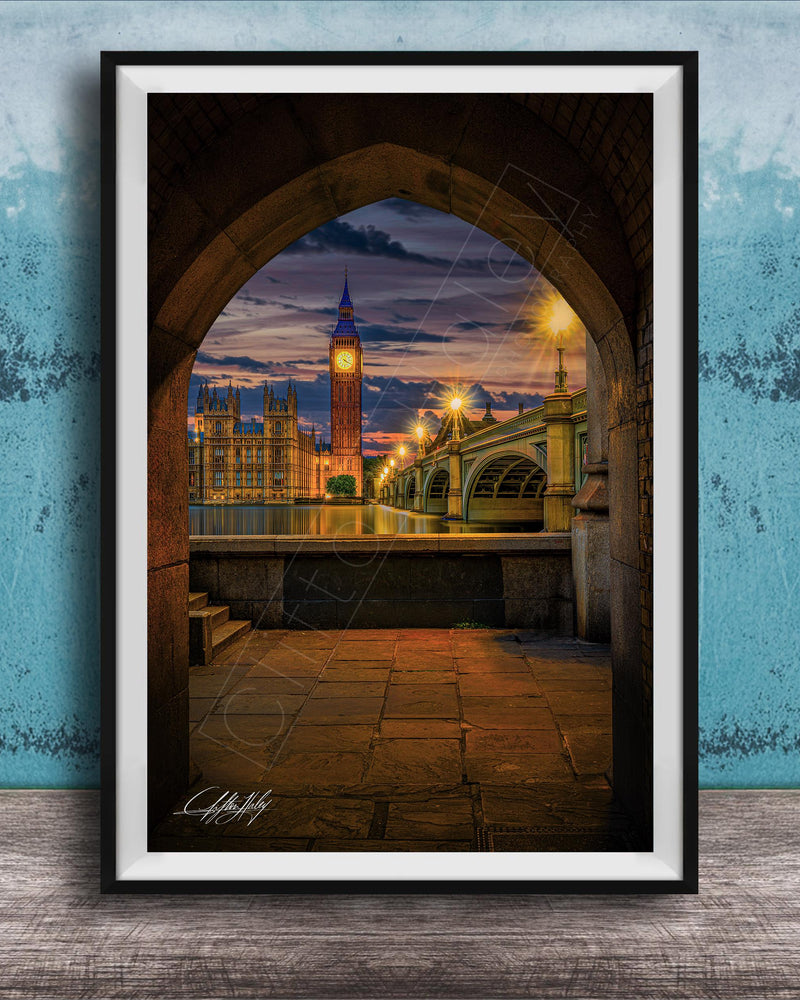 a cityscape at night, with a clock tower and a bridge visible in the background. The clock tower is illuminated, and the bridge is lit up, creating a warm and inviting atmosphere. The scene is framed in a black frame, which contrasts with the blue wall in the background. The image is a collage of multiple images, and the artist's signature is visible in the bottom right corner.
