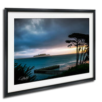 A framed photograph depicting a serene coastal scene with a large cruise ship in the distance, a pier, and a dramatic sky with clouds and a colorful sunset.