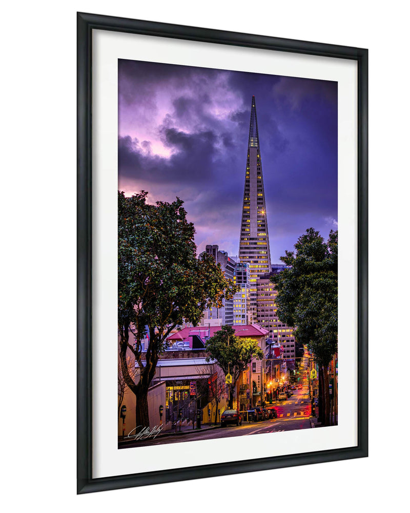 a cityscape at night, with a tall, illuminated skyscraper standing prominently in the background. The city is surrounded by trees and buildings, creating a vibrant and lively atmosphere. The sky above is filled with clouds, adding a dramatic touch to the scene. The image is framed, suggesting it is a piece of art or photography.