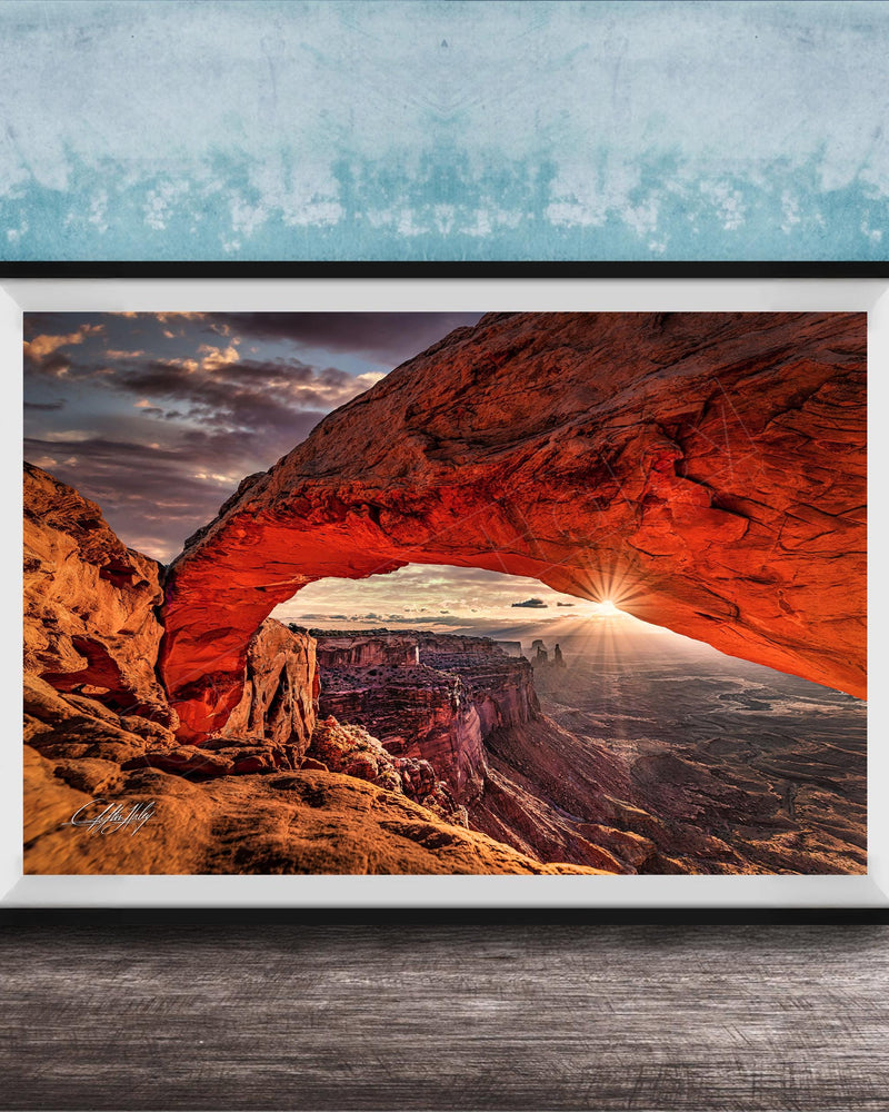 A framed photograph depicting a stunning natural landscape with a large arch formation in the distance, set against a backdrop of a cloudy sky and a sunset.