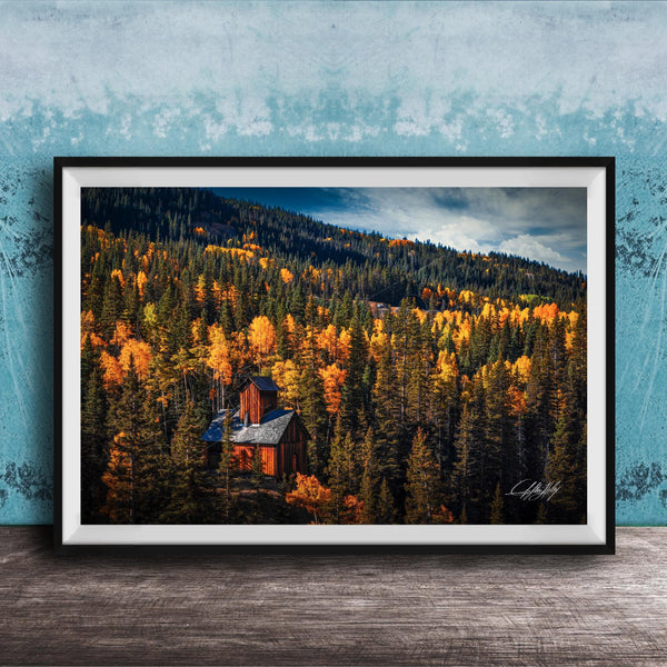 A framed photograph of a cabin nestled in a forest of trees with vibrant autumn foliage.