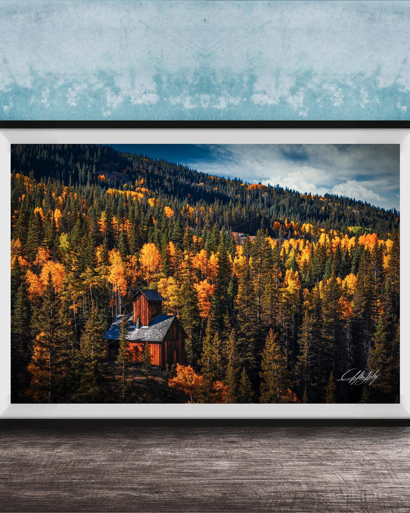 A framed photograph of a cabin nestled in a forest of trees with vibrant autumn foliage.