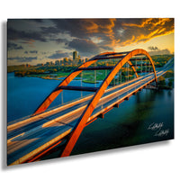 a large, curved bridge spanning across a body of water, with a city skyline visible in the background. The bridge is painted in a striking orange color and has a distinctive arch design. The sky above the scene is filled with vibrant colors, suggesting either sunrise or sunset. The image appears to be a collage of multiple images, with the focus on the bridge and the cityscape.
