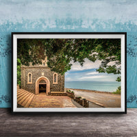 A framed photograph depicting a stone building with arched windows and a stone walkway leading to a beach with a blue sky and trees in the background.