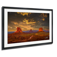 A framed photograph depicting the iconic rock formations of Monument Valley, with the sun setting behind the mountains and a dramatic sky.