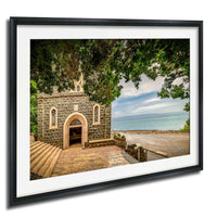 A stone church with a brick walkway and a stone wall, surrounded by trees and overlooking a body of water.