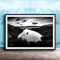 A black and white photograph of a serene lake surrounded by a forested landscape, with a cloudy sky above.