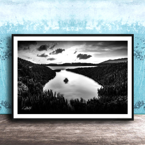 A black and white photograph of a serene lake surrounded by a forested landscape, with a cloudy sky above.