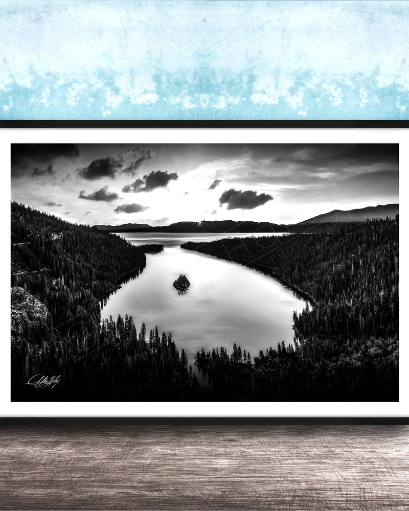 A black and white photograph of a serene lake surrounded by a forested landscape, with a cloudy sky above.