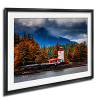 A framed photograph depicting a lighthouse surrounded by a forest with autumn-colored trees and a cloudy sky in the background.