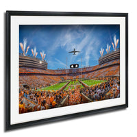 A framed photograph depicting a crowded football stadium during a game, with a plane flying overhead and colorful fireworks lighting up the sky.