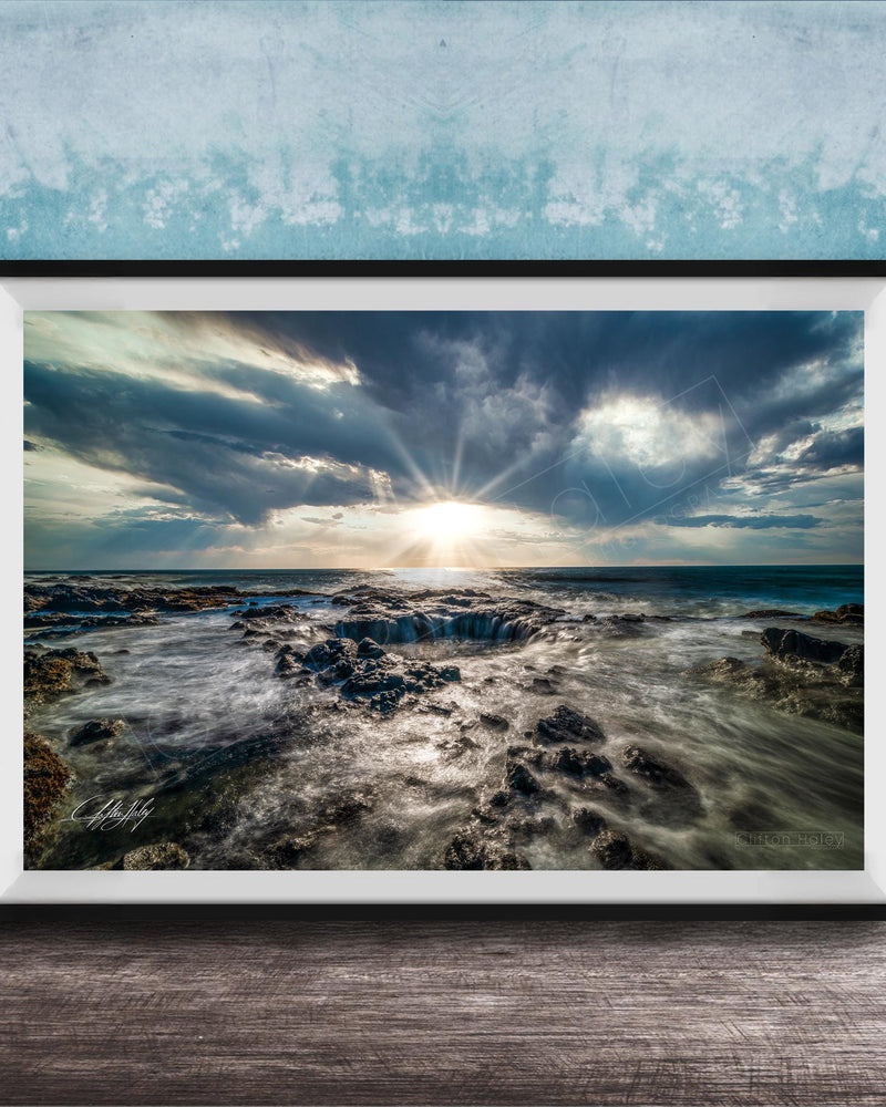 The image is a framed photograph of a rocky coastline with crashing waves, set against a backdrop of a cloudy sky and the sun peeking through the clouds.