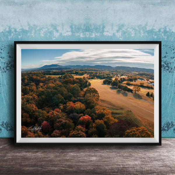 The image is a framed aerial view of a scenic landscape with a mix of trees, fields, and mountains in autumn colors.