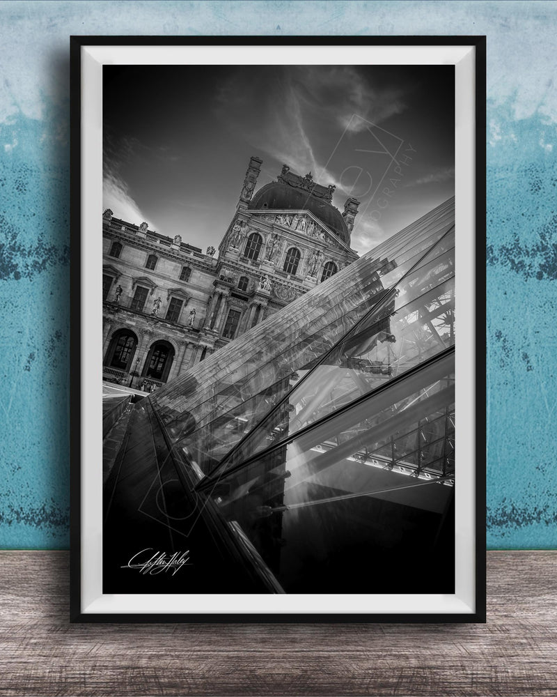 A black and white photograph of the Louvre Museum in Paris, France, with a modern glass structure in the foreground.