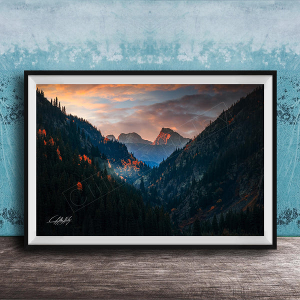 The image is a framed photograph of a mountainous landscape with a sunset sky, featuring a valley with trees and mountains in the background.