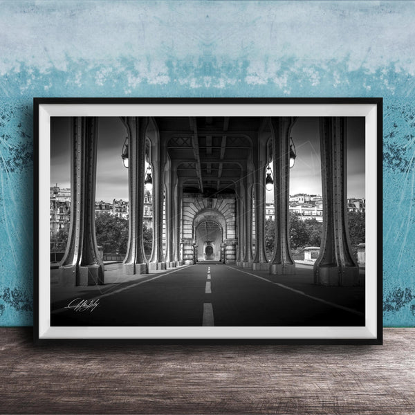 A black and white photograph of a long, narrow bridge with tall columns, leading to a central archway. The bridge is surrounded by trees and buildings, creating a serene and picturesque scene.