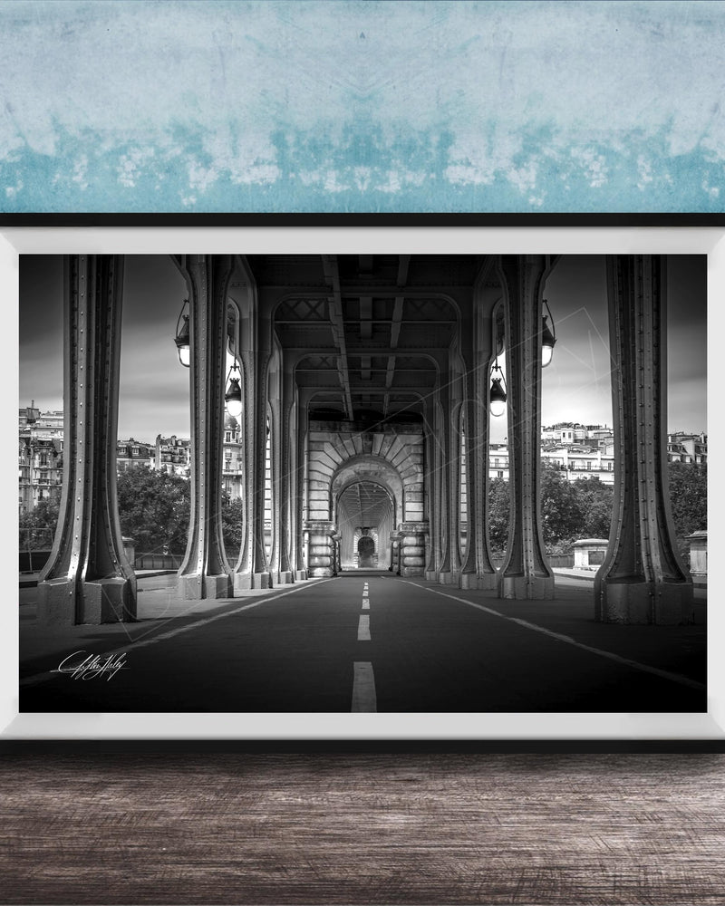 A black and white photograph of a long, narrow bridge with tall columns, leading to a central archway. The bridge is surrounded by trees and buildings, creating a serene and picturesque scene.