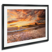 A framed photograph depicting a serene beach scene at sunset, with a wooden pier extending into the water and the sky filled with vibrant orange and pink hues.