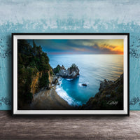 A framed photograph depicting a scenic coastal landscape with a rocky shoreline, a sandy beach, and a body of water with a sunset sky in the background.
