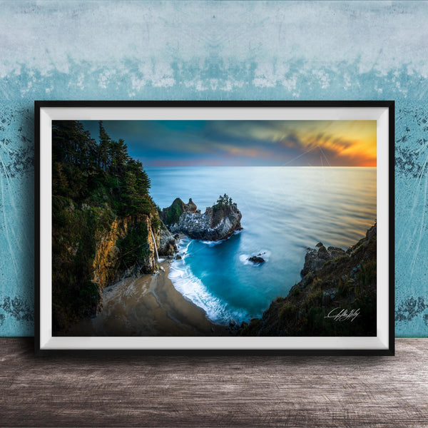 A framed photograph depicting a scenic coastal landscape with a rocky shoreline, a sandy beach, and a body of water with a sunset sky in the background.