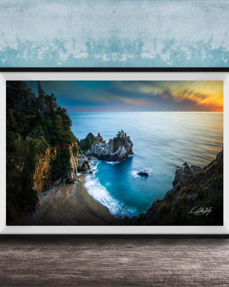 A framed photograph depicting a scenic coastal landscape with a rocky shoreline, a sandy beach, and a body of water with a sunset sky in the background.