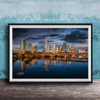 A framed photograph of a city skyline at sunset, with the buildings reflected in the water below.