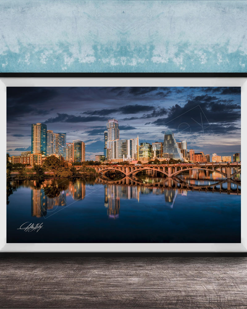 A framed photograph of a city skyline at sunset, with the buildings reflected in the water below.
