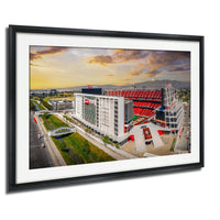 The image is a framed picture of a large stadium with a red roof, surrounded by a cityscape with buildings and a bridge.