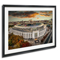 The image is a framed photograph of a large stadium, likely a sports stadium, with a city skyline in the background. The photograph is taken from an aerial perspective, showcasing the stadium and surrounding urban landscape.