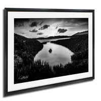 A black and white landscape photograph depicting a serene lake surrounded by a forested area, with mountains visible in the distance under a cloudy sky.