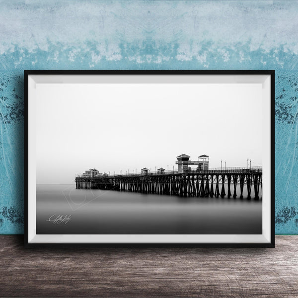 A black and white photograph of a long pier extending into the distance, with a calm body of water below.