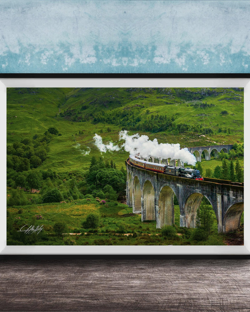 A framed photograph depicting a steam train traveling on a stone bridge over a lush, green valley.