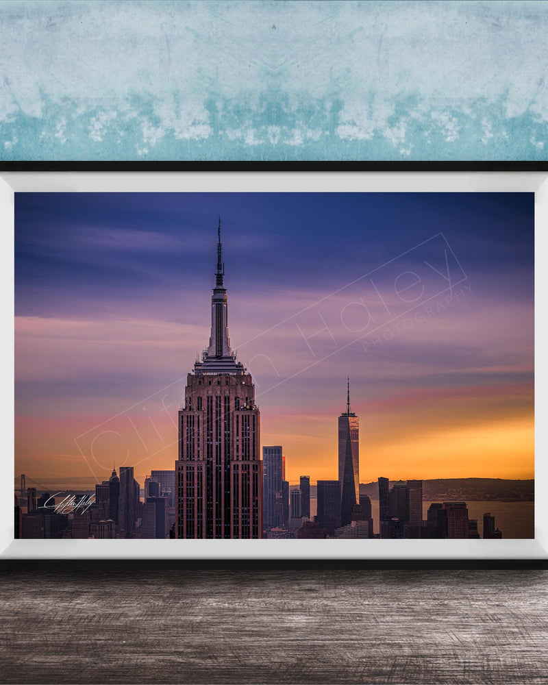 A framed photograph of the New York City skyline at sunset, with the Empire State Building prominently visible in the center of the frame.