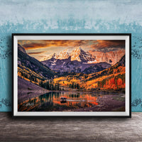 a breathtaking mountain landscape with a lake in the foreground, surrounded by vibrant autumn foliage. The mountains are covered in snow, creating a stunning contrast against the colorful sky. The scene is framed in a black frame and displayed on a wooden surface, adding to the rustic charm of the setting.