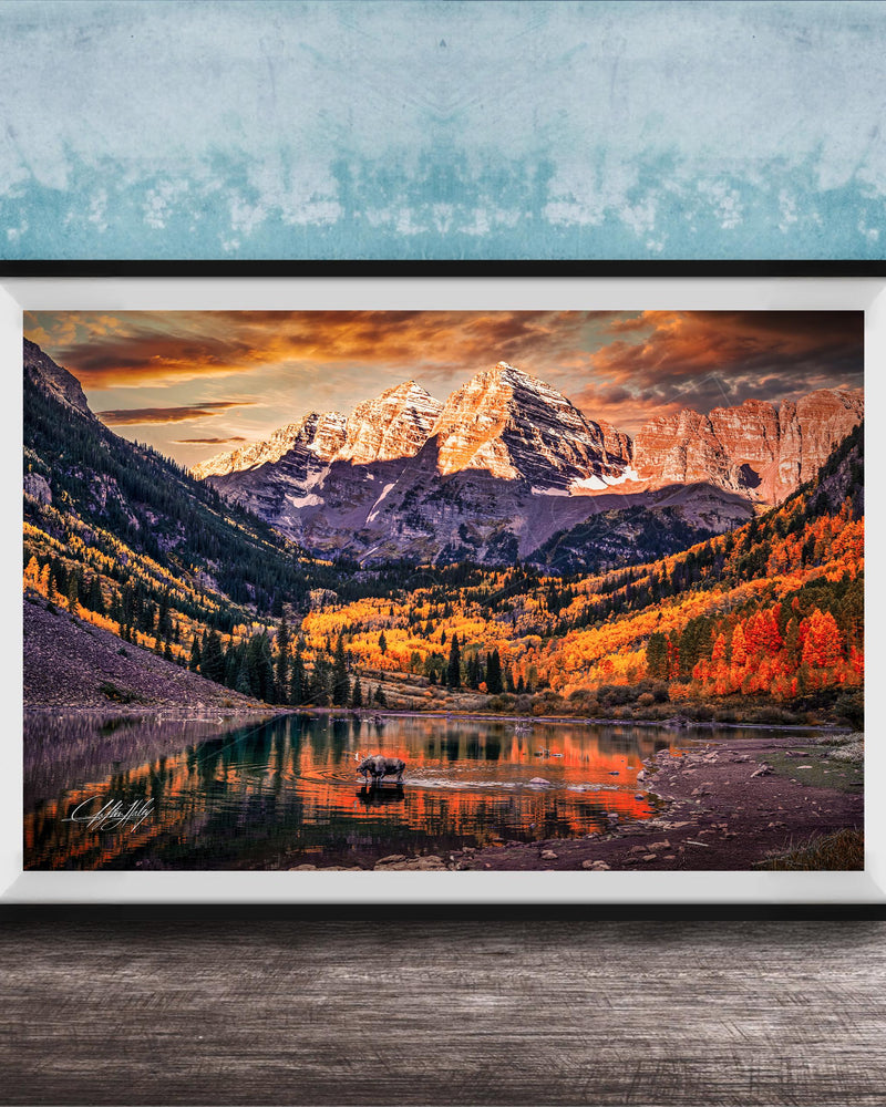 a breathtaking mountain landscape with a lake in the foreground, surrounded by vibrant autumn foliage. The mountains are covered in snow, creating a stunning contrast against the colorful sky. The scene is framed in a black frame and displayed on a wooden surface, adding to the rustic charm of the setting.