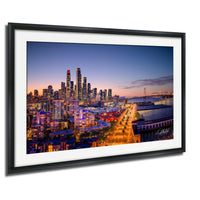 The image is a framed photograph of a city skyline at dusk, with the city lights illuminating the buildings and the surrounding area.