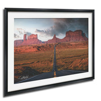 a long, straight road cutting through a vast, arid landscape with towering red rock formations in the background.