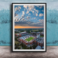 The image is an aerial view of a large sports stadium with a football field, surrounded by buildings and a cloudy sky.