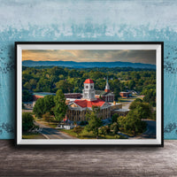 A framed aerial view of a historic building with a red roof, surrounded by lush greenery and mountains in the distance.