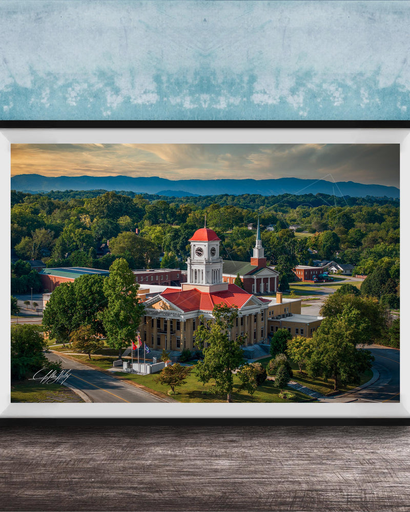 A framed aerial view of a historic building with a red roof, surrounded by lush greenery and mountains in the distance.
