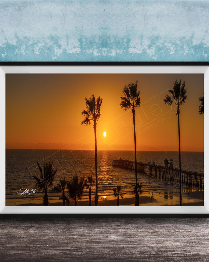 A framed photograph of a serene beach scene at sunset, with palm trees silhouetted against the warm orange sky.