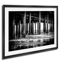 A black and white photograph of a pier with numerous wooden columns and beams, partially submerged in water.