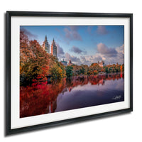 a serene cityscape with a calm lake reflecting the surrounding buildings and trees. The buildings have a mix of architectural styles, and the trees are adorned with autumn foliage, creating a picturesque scene.