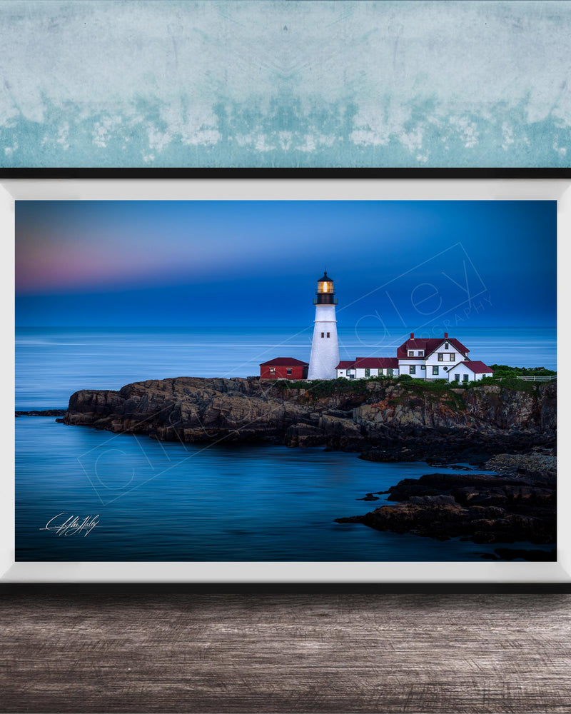 A framed photograph of a lighthouse on a rocky coastline, with the ocean and a colorful sky in the background.