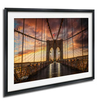 A framed photograph of the Brooklyn Bridge at sunset, with the iconic stone towers and suspension cables illuminated against a vibrant sky.